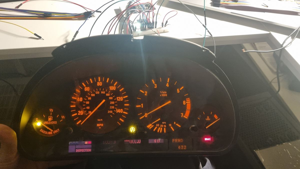 A BMW E39 instrument cluster lit up on a desk, with the speedometer, rev counter and fuel gauge visible, some warning lights on and a breadboard with jumper wires above it.