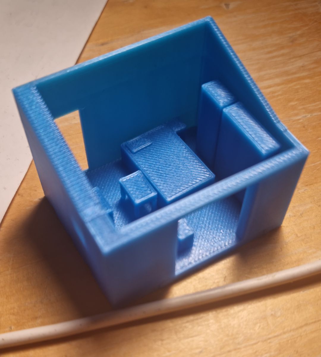 Blue 3D-printed model of a room on a wooden table, open at the top, with simple blocks for the furniture inside and a white cable lying in front of it